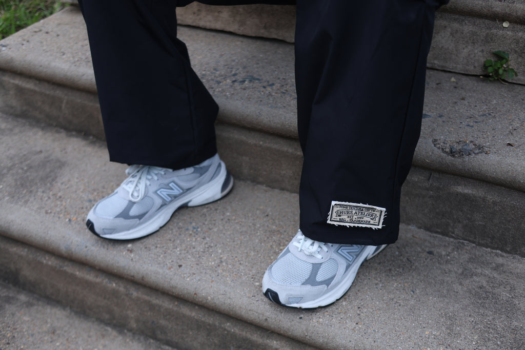 Close-up of black Hues Atelier Track Pants with woven ankle patch worn with white sneakers
