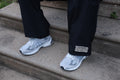 Close-up of black Hues Atelier Track Pants with woven ankle patch worn with white sneakers