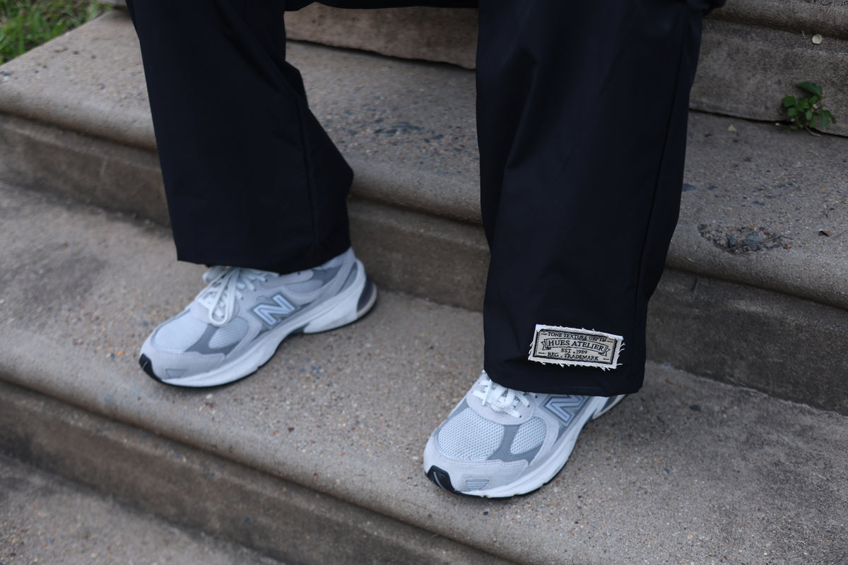 Close-up of black Hues Atelier Track Pants with woven ankle patch worn with white sneakers