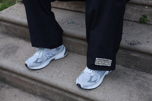 Close-up of black Hues Atelier Track Pants with woven ankle patch worn with white sneakers