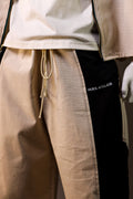 Close-up of beige pants with a drawstring and black patch displaying 'HUES ATELIER' on a neutral background.