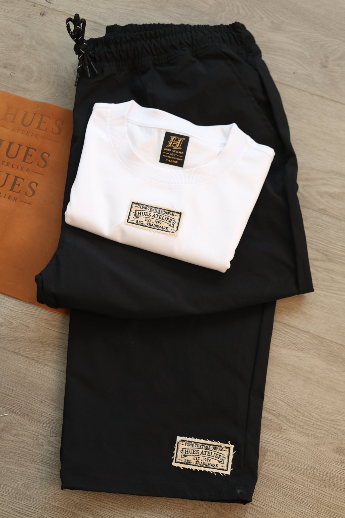 Folded black Hues Atelier Track Pants showing signature woven brand patch