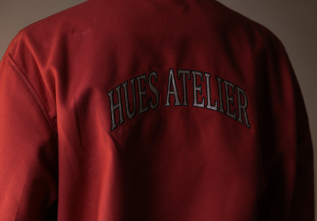 Red jacket with 'HUES ATELIER' text on the back against a neutral background