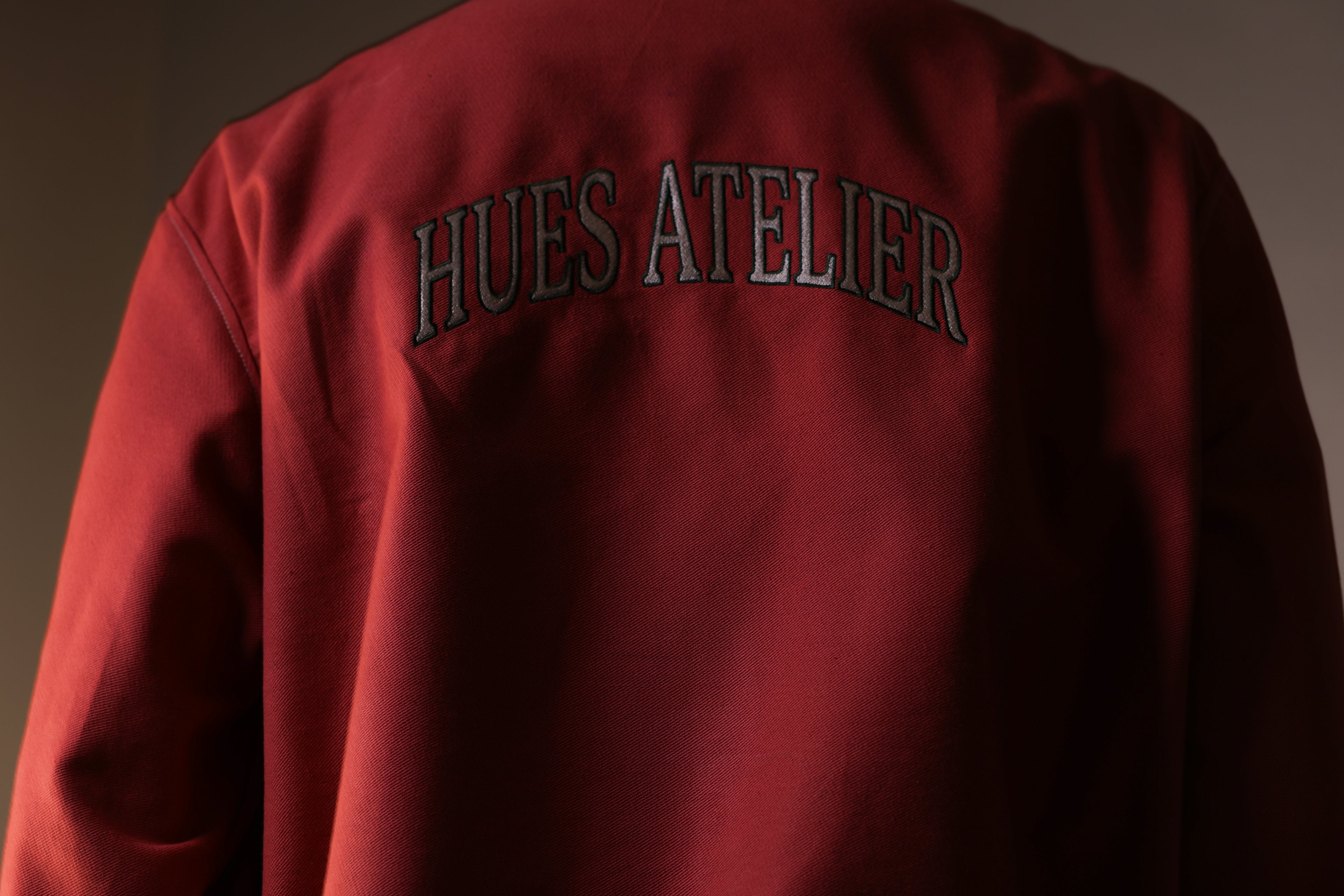 Red jacket with 'HUES ATELIER' text on the back against a dark background