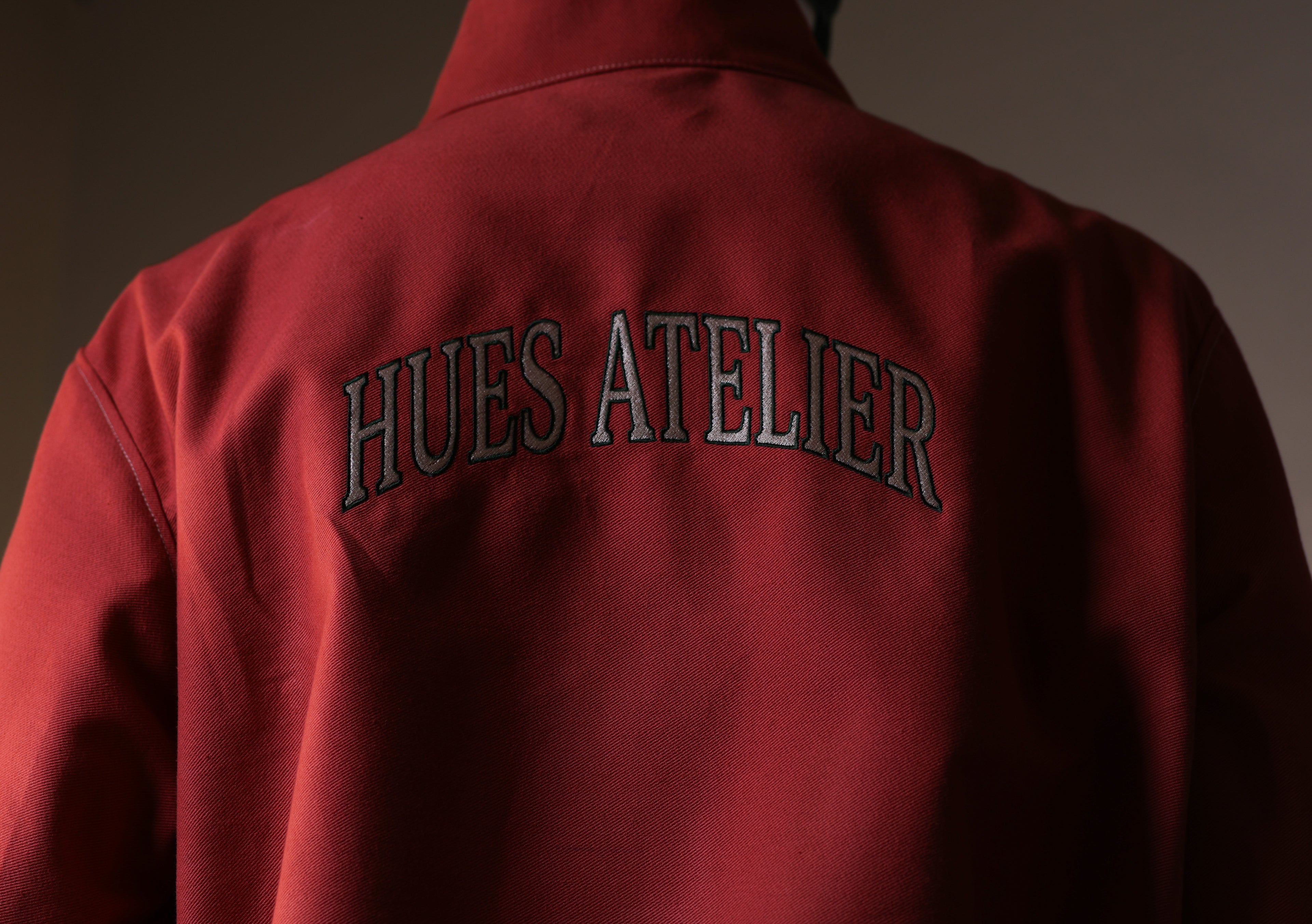 Red jacket with 'HUES ATELIER' text on the back against a dark background