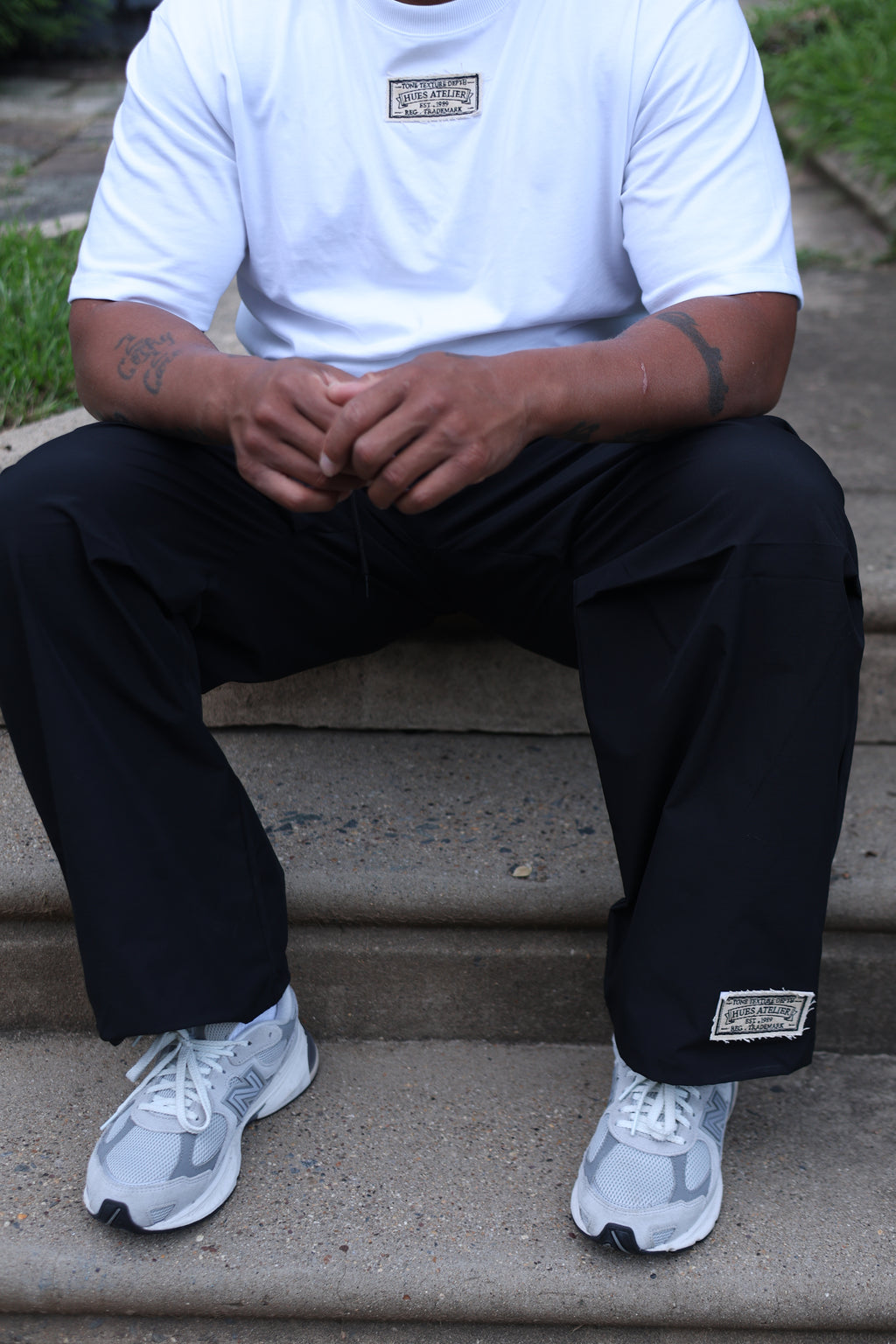 Seated model wearing black Hues Atelier Atelier Track Pants with a white T-shirt