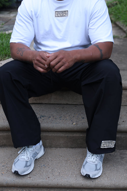Seated model wearing black Hues Atelier Atelier Track Pants with a white T-shirt